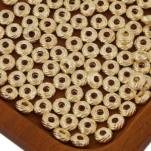 May include: A collection of gold-coloured beads with a spiral design. Each bead has a central hole. The beads are arranged on a wooden surface. These beads are suitable for jewellery making and various craft projects. The beads are approximately 1 cm in diameter.