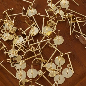 May include: A pile of gold-toned earring posts with loops. The posts are made of metal and have a round base. The loops are small and are located at the top of the posts.