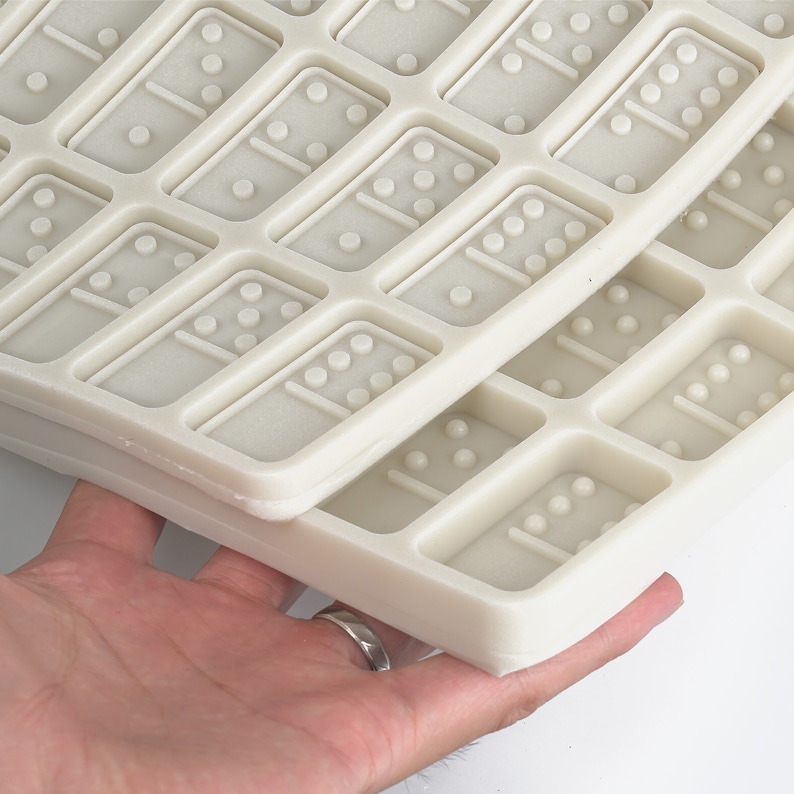 May include: Two white silicone molds for making dominoes. The molds are stacked on top of each other and show the raised details of the dominoes.