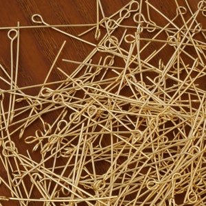 May include: A large pile of gold-colored head pins with a loop at one end. The pins are scattered randomly on a wooden surface.