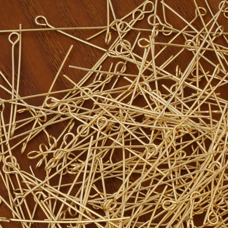 May include: A large pile of gold-colored head pins with a loop at one end. The pins are scattered randomly on a wooden surface.