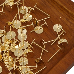 May include: A close-up image of a pile of gold-coloured earring posts. The posts are round and have a flat back. They are scattered on a wooden surface.
