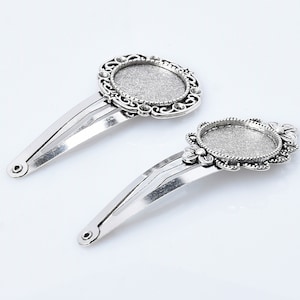 May include: Two silver hair clips with ornate, round, silver-toned metal frames. The frames have a smooth, circular, clear glass or plastic insert in the centre.