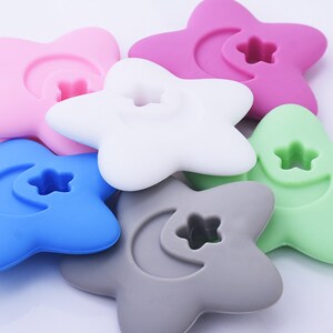 May include: A set of six silicone star-shaped teethers in various colors, including pink, white, blue, gray, and green. Each teether features a crescent moon and a star cutout.
