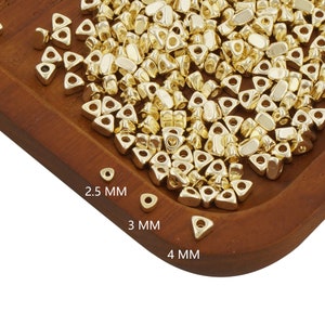 May include: A large pile of gold-colored triangle-shaped beads with a hole in the center. The beads are arranged on a wooden surface. The text "2.5 MM", "3 MM", and "4 MM" is printed on the surface.