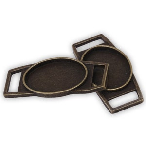 May include: Two antique gold-toned metal oval-shaped blanks with a loop on each side. The blanks are designed for adding a cameo or other embellishment.