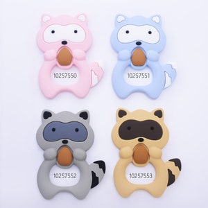 May include: Four colorful silicone baby teethers shaped like raccoons. The teethers are in pink, blue, gray, and tan, each with a brown center and a white belly. Each teether has a unique number printed on it.