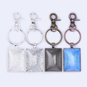 Peut inclure: Quatre porte-clés avec des inserts carrés en verre ou en métal. Deux porte-clés sont argentés avec un insert argenté, un est bronze avec un insert bronze et un est bronze avec un insert en verre bleu. Chaque porte-clés a un anneau brisé et un fermoir à homard.
