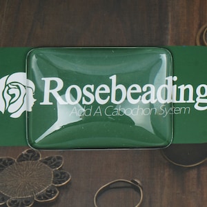 May include: A green, rectangular, clear plastic tray with the text "Rosebeading Add A Cabochon System" printed in white.