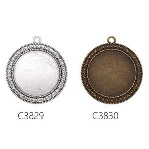 May include: Two round metal pendant blanks with a decorative border. The blank on the left is silver and the blank on the right is bronze. Both blanks have a hole at the top for hanging.
