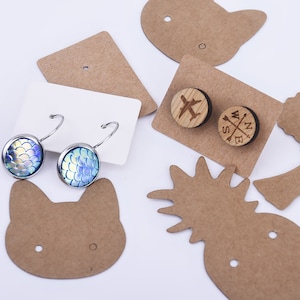 May include: A set of earrings with a variety of designs. The earrings are displayed on brown paper cards with different shapes, including a cat, a pineapple, and an airplane. The earrings include a pair of silver hoop earrings with blue and green iridescent mermaid scale designs, a pair of wooden earrings with a compass design, and a pair of wooden earrings with an airplane design.