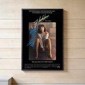 May include: A framed movie poster for "Flashdance" featuring a woman in a gray sweater and red heels. The title is in a stylized font with blue accents. The poster has the tagline "Take your passion and make it happen!"