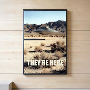 Könnte beinhalten: Gerahmter Druck mit einer metallischen UFO, die über einer Wüstenlandschaft mit Bergen im Hintergrund schwebt. Der Text "THEY'RE HERE" ist in weißen Blockbuchstaben am unteren Bildrand zu sehen.