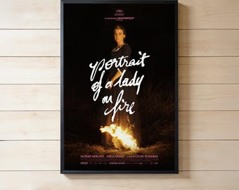 Pode incluir: Um cartaz de filme emoldurado de Portrait of a Lady on Fire mostrando uma pessoa com um vestido escuro num campo com chamas na bainha. O título manuscrito branco está centrado, com os nomes do elenco e do realizador na parte inferior.