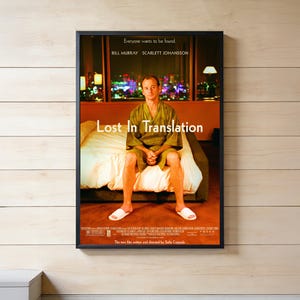 以下が含まれることがあります： ホテルの一室のベッドに座るビル・マーレイが描かれた映画『ロスト・イン・トランスレーション』の額入りポスター。ポスターには白い文字で映画タイトル、ビル・マーレイとスカーレット・ヨハンソンの名前、そしてキャッチコピーの「Everyone wants to be found.」が記載されています。