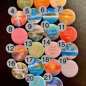 May include: A collection of 21 round buttons with the text "Eternal Happiness 2026". The buttons feature various designs, including beach scenes, sunsets, and solid colors like pink, orange, and blue. The buttons are approximately 1.5 inches in diameter.