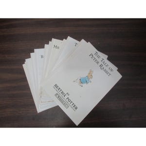 Peut inclure: Une pile de livres pour enfants anciens, avec le titre "The Tale of Peter Rabbit" de Beatrix Potter. La couverture montre un lapin en veste bleue. Les livres sont disposés en éventail sur une surface en bois sombre.
