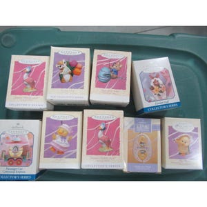 May include: A collection of Hallmark Keepsake ornaments in their original boxes. The ornaments feature various characters and designs, including animals and a train. The boxes are primarily white, pink, and purple.