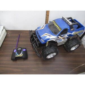 Pode incluir: Um caminhão de brinquedo Ford azul e branco com pneus pretos grandes e um controle remoto preto. O caminhão tem as palavras "Street Beast" e "Ford" impressas. O controle remoto é preto com detalhes roxos.