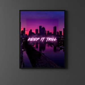 May include: A framed art print depicting a city skyline against a purple and pink sunset. The neon text "KEEP IT TRILL" is displayed across the image. The artwork is mounted on a dark grey wall.