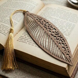 Könnte beinhalten: Ein hölzerner Lesezeichen in Form eines Blattes mit aufwendigen Ausschnitten liegt auf den Seiten eines offenen Buches. Eine goldene Quaste hängt am oberen Ende des Lesezeichens. Eine Brille und eine Tasse Tee sind im Hintergrund zu sehen.