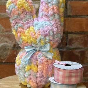 May include: A pastel-colored, plush bunny with a light blue satin bow. The bunny is made of soft, textured yarn in shades of yellow, pink, blue, and green. Two spools of gingham ribbon in pink and blue are also visible.