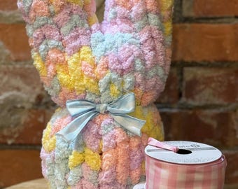 May include: A pastel-colored, plush bunny with a light blue satin bow. The bunny is made of soft, textured yarn in shades of yellow, pink, blue, and green. Two spools of gingham ribbon in pink and blue are also visible.
