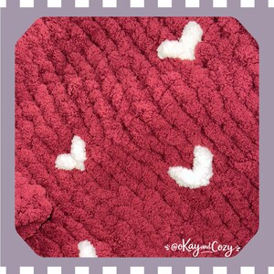 May include: A burgundy chunky knit blanket with white heart-shaped accents. The blanket has a soft, textured appearance and is suitable for home decor. The text "@Kay and Cozy" is in the bottom right corner.