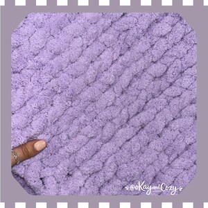 May include: Close-up of a soft, lavender-colored knitted blanket. The chunky knit texture is visible, with diagonal rows of thick yarn. The blanket appears cozy and warm, perfect for snuggling. The text "@Kay and Cozy" is in the bottom right corner.