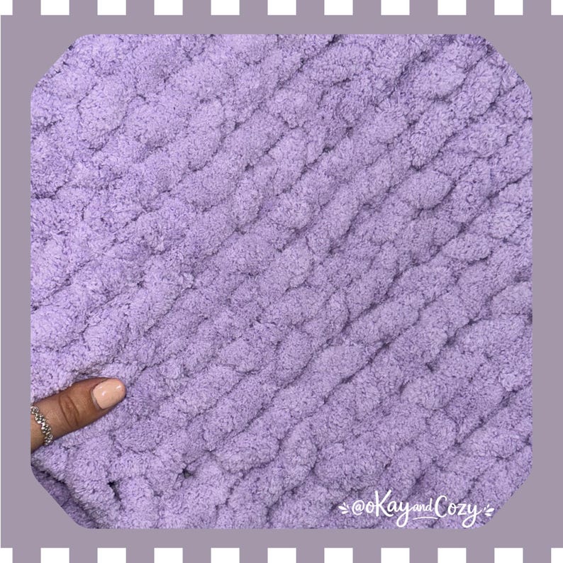 May include: Close-up of a soft, lavender-colored knitted blanket. The chunky knit texture is visible, with diagonal rows of thick yarn. The blanket appears cozy and warm, perfect for snuggling. The text "@Kay and Cozy" is in the bottom right corner.