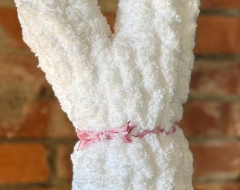 May include: A fluffy white towel shaped like a bunny, with two upright ears and a round body. A pink ribbon is tied around the middle. The background is a blurred brick wall and a wooden surface.