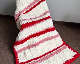 May include: A handmade, chunky knit blanket in white, red, and pink. The blanket features thick, horizontal stripes of red and pink against a white background. The edges are trimmed with a red border. A small red heart is visible on the top edge.