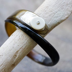 Könnte beinhalten: Ein schwarzes und braunes Armreif-Armband mit einem weißen und silbernen Akzent. Der Akzent hat einen kleinen, klaren Stein.