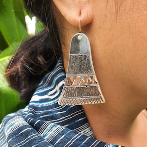 May include: Silver dangle earrings with a geometric design. The earrings are shaped like a triangle with a pointed top and a rounded bottom. The design features a series of lines and triangles that create a geometric pattern.