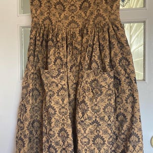 May include: A corduroy dress in tan with a navy floral pattern, featuring a square neckline, short sleeves, and two front pockets. The dress has a fitted bodice and a flared skirt. It is hanging on a wooden hanger.