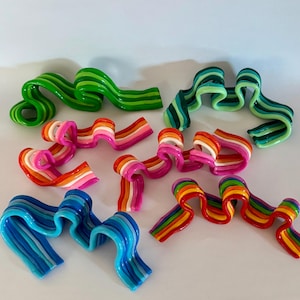 Puede incluir: Una colección de esculturas coloridas en forma de cinta en varios tonos, incluyendo verde, azul, rosa y patrones de arcoíris. Cada pieza tiene un acabado brillante y un diseño ondulado, creando una exhibición lúdica y artística.