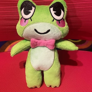 May include: A plush frog toy featuring bright green fabric, a light green belly, and a pink bowtie. The frog has large embroidered black eyes with white highlights and pink circular patches on its cheeks, with a simple black stitched mouth.