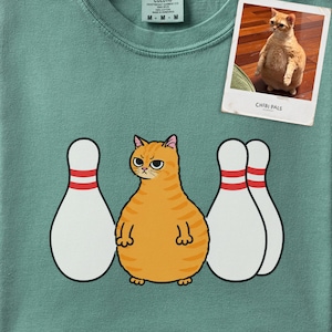 Può includere: Una t-shirt verde salvia con la grafica di un gatto soriano arancione in piedi tra tre birilli da bowling bianchi con strisce rosse. In alto a destra è presente una stampa fotografica rettangolare di un gatto arancione con il testo CHIBI PALS.