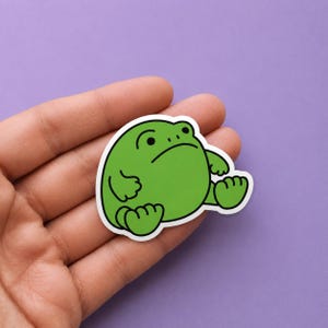Puede incluir: Una pegatina troquelada que muestra una rana redonda de color verde brillante con un contorno negro sencillo. La rana aparece en posición sentada con una expresión facial neutra, caracterizada por dos pequeños puntos negros como ojos.