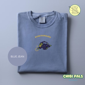 May include: A blue jean Comfort Colors t-shirt with the text "BOOM SHAKALAKA" above an embroidered cartoon frog wearing a wizard robe. The frog is holding a yellow lightning bolt. The shirt has a crew neck and is folded.