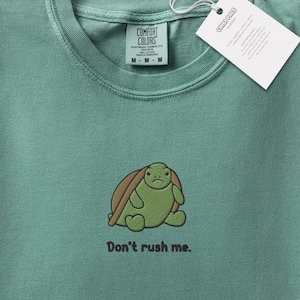 Könnte beinhalten: Ein salbeigrünes T-Shirt mit einer Stickerei einer kleinen, mürrisch aussehenden Schildkröte mit braunem Panzer. Unter der Schildkröte steht der Text "Don't rush me.". Das Shirt hat ein Comfort Colors Markenetikett und ein weißes Chibi Pals Etikett.