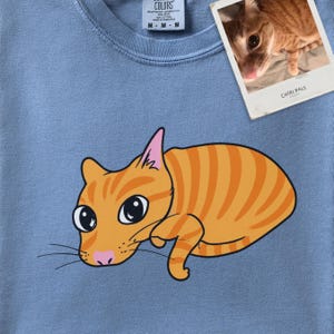 May include: A light blue t-shirt featuring a graphic illustration of an orange tabby cat with large black eyes and pink ears. A small white rectangular photo print of the same orange cat is attached to the top right, labeled Chibi Pals.