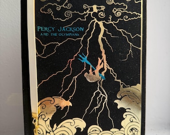 May include: A hardcover book titled "Percy Jackson and the Olympians" with a black cover and gold foil accents. The cover features a lightning bolt design and a figure holding a trident. A white ribbon bookmark with a gold charm is attached.