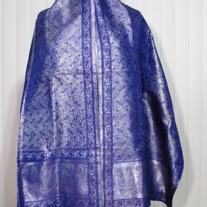 May include: A rectangular shawl in a deep royal blue color featuring an intricate silver metallic paisley pattern throughout. The garment includes a wide decorative border with a repeating paisley motif and finished with silver fringe at the bottom.