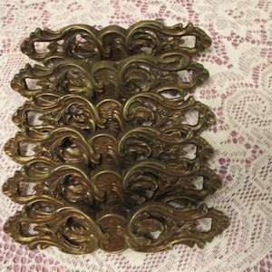 May include: A set of six matching brass drawer pulls featuring an ornate, symmetrical scrollwork design with intricate leaf-like detailing and a central textured oval element. Each handle has a curved, elongated shape with a metallic finish.