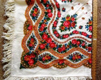 May include: A square off-white shawl featuring a vibrant floral pattern of red roses and blue flowers with orange wavy borders and long fringe edges. A wooden ruler placed along the side shows the fabric measures approximately 36 inches in length.