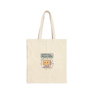May include: Cream-colored tote bag featuring a retro computer graphic with a smiling face and the text "Buffering... Please Wait." The design includes hearts, stars, and other decorative elements. The bag has long handles.