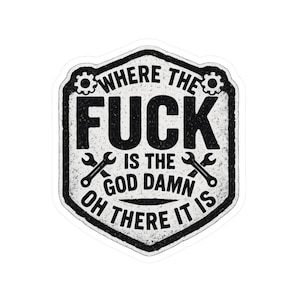 Puede incluir: Una pegatina en blanco y negro con el texto "WHERE THE FUCK IS THE GOD DAMN OH THERE IT IS" en una fuente desgastada. La pegatina tiene forma de escudo con engranajes y llaves inglesas.