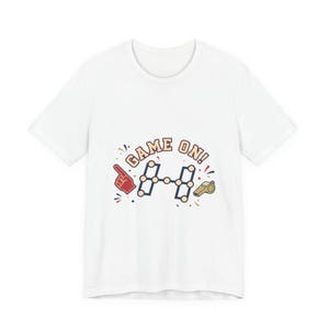 Puede incluir: Camiseta blanca con un gráfico colorido que dice "GAME ON!". El diseño incluye un dedo de espuma rojo, una pesa azul y blanca y un silbato dorado. Un diseño divertido y deportivo.