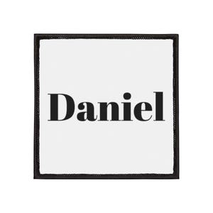 Peut inclure: Écusson carré blanc avec le nom "Daniel" en lettres noires grasses. Le patch est bordé d'un liseré noir avec un aspect cousu.